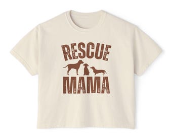 May include: Cream-colored t-shirt with the words "RESCUE MAMA" in distressed brown lettering. The design includes silhouettes of three dogs: a standing dog, a sitting dog, and a dachshund.