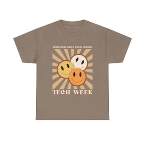May include: A taupe short-sleeve t-shirt featuring a graphic of three smiling faces in yellow, orange, and white. Behind the faces is a tan sunburst pattern. The text above reads SORRY FOR WHAT I SAID DURING and below reads TECH WEEK.