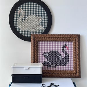 May include: Two framed cross-stitch swan designs, one round and one square, with gingham backgrounds. A white and a navy blue storage box are stacked on a white surface. A pair of small scissors and thread are also on the surface.
