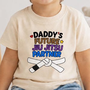 May include: A cream-coloured short-sleeve t-shirt featuring the text "DADDY'S FUTURE JIU JITSU PARTNER" in black, brown, purple, and blue, accompanied by a small red heart, a yellow star, and a graphic of a white martial arts belt.