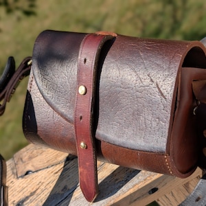 Peut inclure: Sac en cuir marron artisanal avec fermeture à rabat et bandoulière réglable. Le sac présente une quincaillerie en laiton et une finition texturée. Le design comprend une sangle avant fixée avec des rivets en laiton et une sangle latérale avec une boucle en laiton.