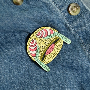 May include: A yellow, green, and pink enamel pin with a detailed, abstract design. The pin is shaped like a face with a large eye and a mouth.