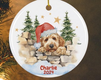 May include: A round Christmas ornament featuring a watercolor illustration of a dog wearing a Santa hat, peeking over a stone wall. The ornament has the name "Charlie" and the year "2024" printed on it. Gold string for hanging.
