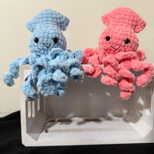 Könnte beinhalten: Zwei gehäkelte Octopus-Plüschtiere, eines blau und eines rosa, mit lächelnden Gesichtern und lockigen Tentakeln. Die Plüschtiere sitzen in einer weißen Holzkiste.