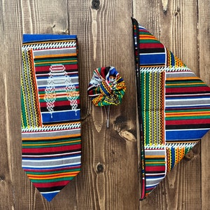 May include: A set of three colorful patterned fabric accessories: a tie, a pocket square, and a bow tie. The tie and pocket square feature a geometric pattern with stripes and squares in shades of red, blue, green, yellow, and black. The bow tie is a smaller version of the pocket square.