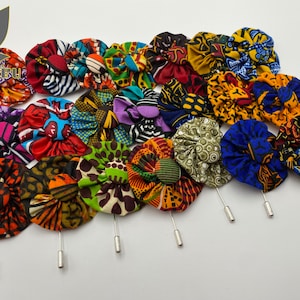 Könnte beinhalten: Eine Sammlung von 17 bunten Stoffblumenbroschen mit Stiften. Die Blumen sind aus verschiedenen afrikanischen Druckstoffen in leuchtenden Farben und Mustern gefertigt.
