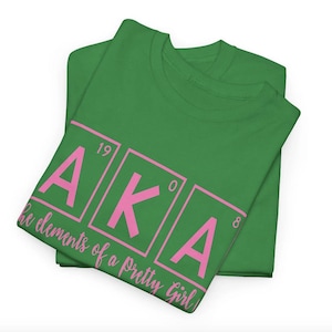 Puede incluir: Camiseta verde con un diseño rosa "AKA". Las letras están en cajas con números encima. El texto debajo dice "The elements of a pretty girl".