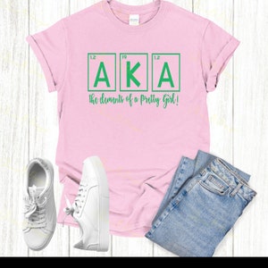 May include: Pink t-shirt with a green graphic of the letters "AKA" in a periodic table style. The text below reads "the elements of a pretty girl!"