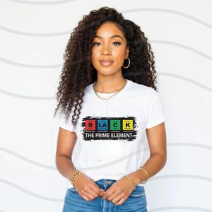 Op de afbeelding: Wit T-shirt met een zwarte afbeelding van het woord "BLACK" gespeld met chemische elementenboxen. De tekst "THE PRIME ELEMENT" staat onder de afbeelding.