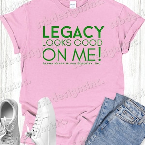 May include: Pink t-shirt with the text "LEGACY LOOKS GOOD ON ME!" in green letters. Below the text is "ALPHA KAPPA ALPHA SORORITY, INC."
