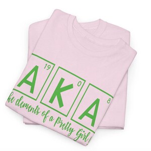May include: Pink t-shirt with the letters AKA in green, styled like a periodic table element. The text below reads "the elements of a pretty girl!"