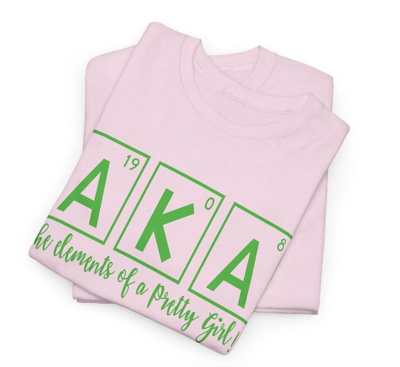 May include: Pink t-shirt with the letters AKA in green, styled like a periodic table element. The text below reads "the elements of a pretty girl!"