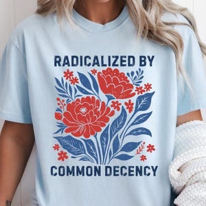 May include: Light blue t-shirt with the text "RADICALIZED BY COMMON DECENCY" in navy blue. The shirt features a floral design with red flowers and blue leaves. The shirt is a casual, comfortable style.