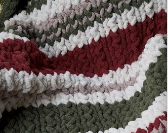 Könnte beinhalten: Eine gefaltete, grob gestrickte Decke mit horizontalen Streifen in Dunkelrot, Olivgrün, Weiß und Grau. Das dicke Garn erzeugt eine strukturierte, gemütliche Optik. Geeignet für Heimdekoration und Komfort, ein warmes Accessoire.