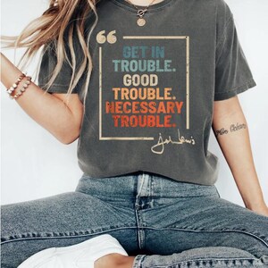 May include: A gray t-shirt with a vintage-style graphic that reads "Get in Trouble. Good Trouble. Necessary Trouble." with a signature below the text. The shirt is worn by a person wearing blue jeans and white sneakers.