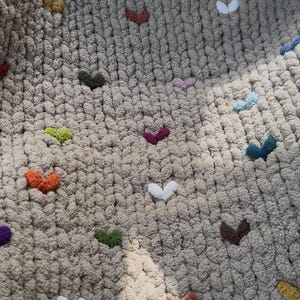 May include: Close-up of a beige chunky knit blanket adorned with colorful heart-shaped accents. The hearts are in various colors, including orange, green, purple, and white, adding a playful touch to the cozy textile.