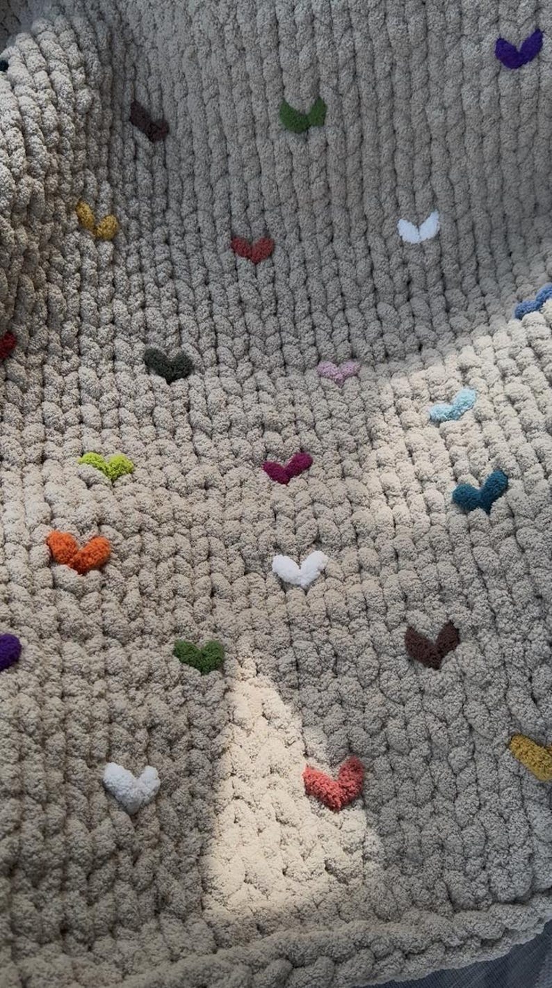 May include: Close-up of a beige chunky knit blanket adorned with colorful heart-shaped accents. The hearts are in various colors, including orange, green, purple, and white, adding a playful touch to the cozy textile.