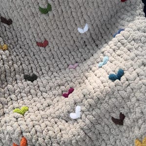 May include: A chunky knit blanket in a neutral beige color, adorned with small, colorful, heart-shaped embellishments. The hearts are in various colors, including white, green, purple, and orange. The blanket appears soft and cozy.