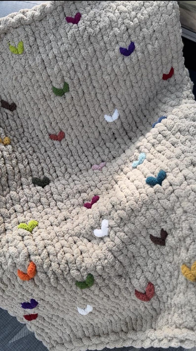 May include: A chunky knit blanket in a neutral beige color, adorned with small, colorful, heart-shaped embellishments. The hearts are in various colors, including white, green, purple, and orange. The blanket appears soft and cozy.