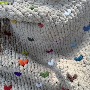 May include: A chunky knit blanket in a neutral beige color, adorned with colorful heart-shaped embellishments. The hearts are in various colors, including red, orange, green, purple, and blue. The blanket has a soft, textured appearance.