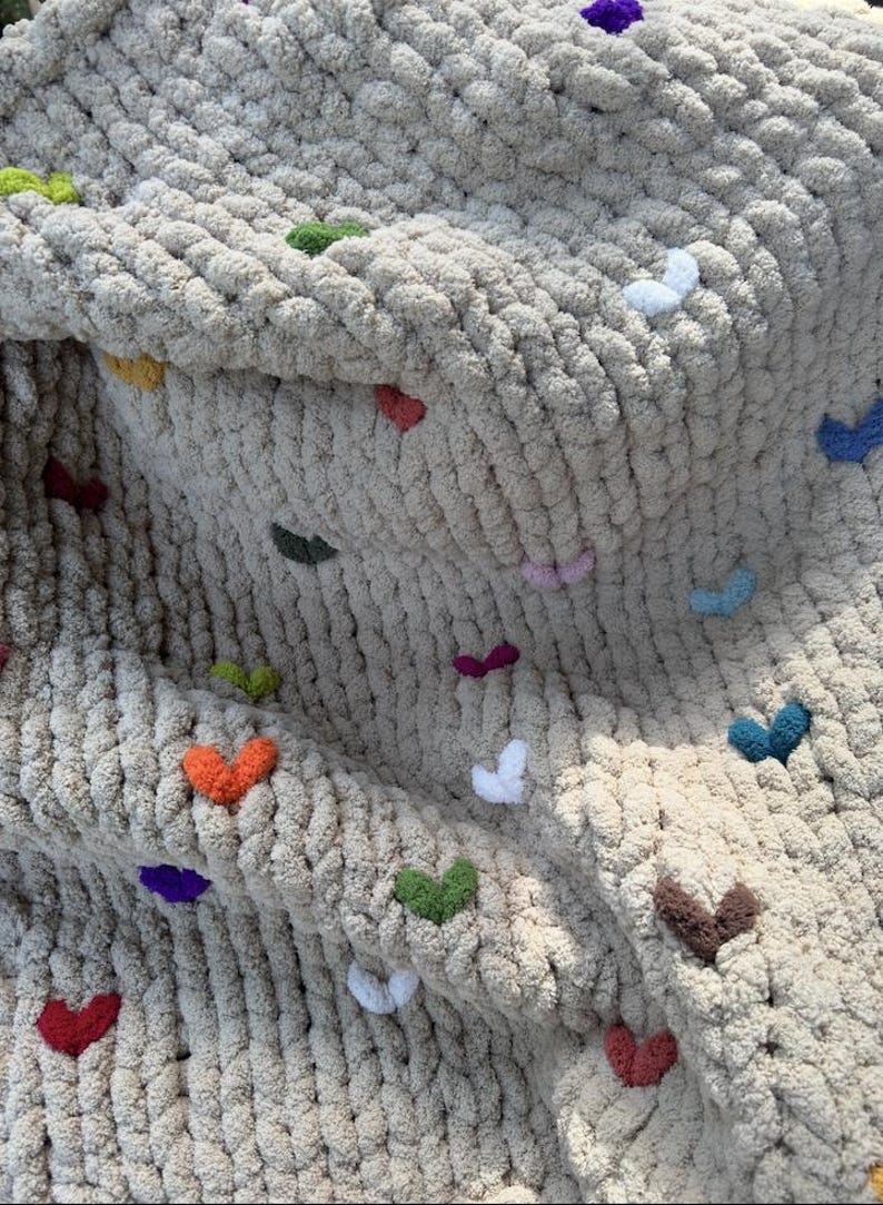 May include: A chunky knit blanket in a neutral beige color, adorned with colorful heart-shaped embellishments. The hearts are in various colors, including red, orange, green, purple, and blue. The blanket has a soft, textured appearance.