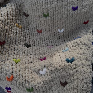 May include: A beige, chunky knit blanket adorned with colorful, embroidered heart-shaped accents. The hearts are in various colors, including red, green, yellow, and purple. The blanket is soft and textured, perfect for home decor.