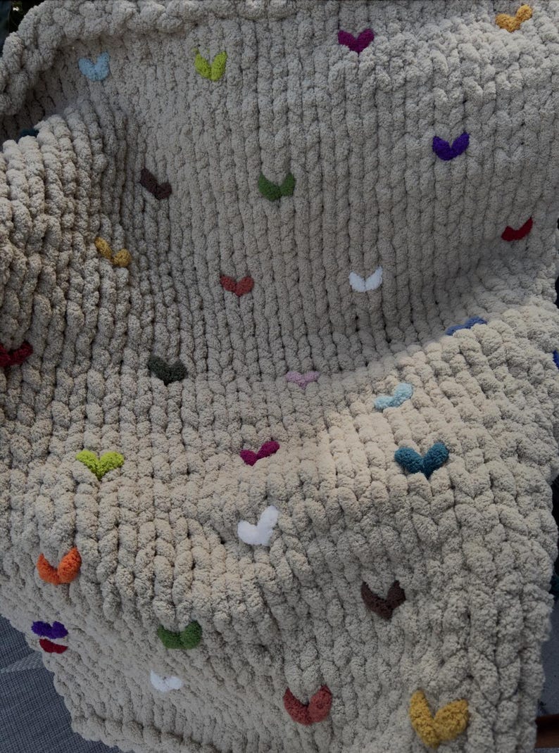 May include: A beige, chunky knit blanket adorned with colorful, embroidered heart-shaped accents. The hearts are in various colors, including red, green, yellow, and purple. The blanket is soft and textured, perfect for home decor.