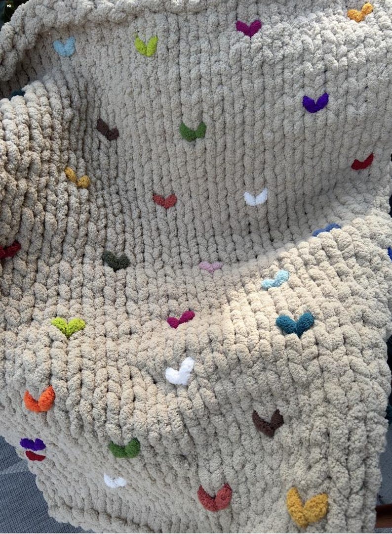 May include: A beige, chunky knit blanket with colorful heart-shaped accents. The hearts are in various colors, including red, green, yellow, purple, and blue. The blanket is soft and textured, perfect for home decor.