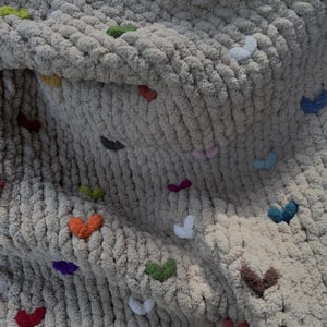May include: A chunky knit blanket in a neutral beige color, adorned with small, colorful heart-shaped accents. The hearts are in various colors, including red, green, orange, and purple, adding a playful touch to the cozy textile.