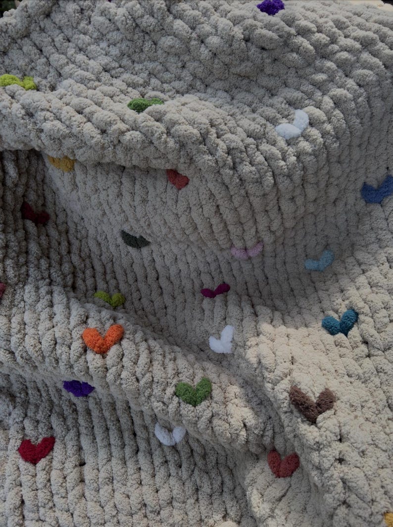 May include: A chunky knit blanket in a neutral beige color, adorned with small, colorful heart-shaped accents. The hearts are in various colors, including red, green, orange, and purple, adding a playful touch to the cozy textile.