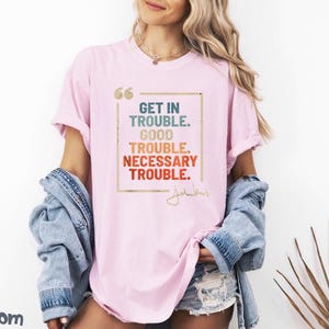 May include: Pink t-shirt with a graphic print that says "Get in Trouble. Good Trouble. Necessary Trouble." with a signature underneath.