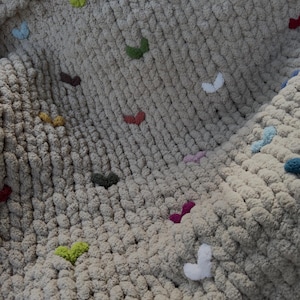 May include: A chunky knit blanket in a neutral beige color, adorned with colorful heart-shaped embellishments. The hearts are in various colors, including red, green, and purple, adding a playful touch to the cozy blanket.