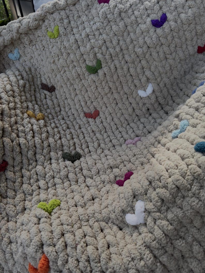 May include: A chunky knit blanket in a neutral beige color, adorned with colorful heart-shaped embellishments. The hearts are in various colors, including red, green, and purple, adding a playful touch to the cozy blanket.