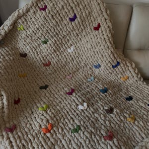 May include: A chunky knit blanket in a neutral beige color, adorned with colorful, embroidered butterfly accents. The butterflies are in various colors, including red, purple, green, and orange. The blanket is draped over a light-colored sofa.