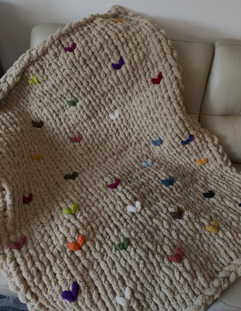 May include: A chunky knit blanket in a neutral beige color, adorned with colorful, embroidered butterfly accents. The butterflies are in various colors, including red, purple, green, and orange. The blanket is draped over a light-colored sofa.