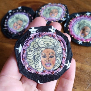 May include: A black and white fabric pin with a pink and white fabric circle in the centre. The circle features a drawing of a woman with blonde hair and a pink and white fabric border with small beads.
