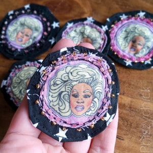 May include: A black fabric circle with white stars and pink and gold beads around the edge. The centre of the circle features a pink fabric background with a drawing of a woman with blonde hair.