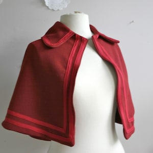 May include: A red cape with a collar and double-stitched trim. The cape is open in the front and has a smooth, slightly textured fabric.