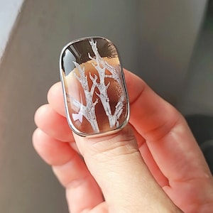 May include: A rectangular silver ring featuring a unique stone with a tree-like pattern. The stone has a brown base with white, branch-like inclusions and a glossy, dark brown top. The ring is held by a hand.