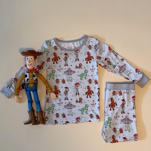 May include: A light blue pajama set with a print of characters from the movie Toy Story. The set includes a long-sleeved top and matching shorts. A Woody action figure is next to the pajama set.