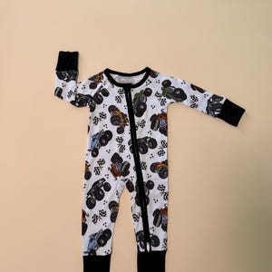 May include: White baby sleeper with a colorful monster truck pattern. The one-piece outfit features a black zipper, cuffs, and collar. The design includes monster trucks and checkered flags.