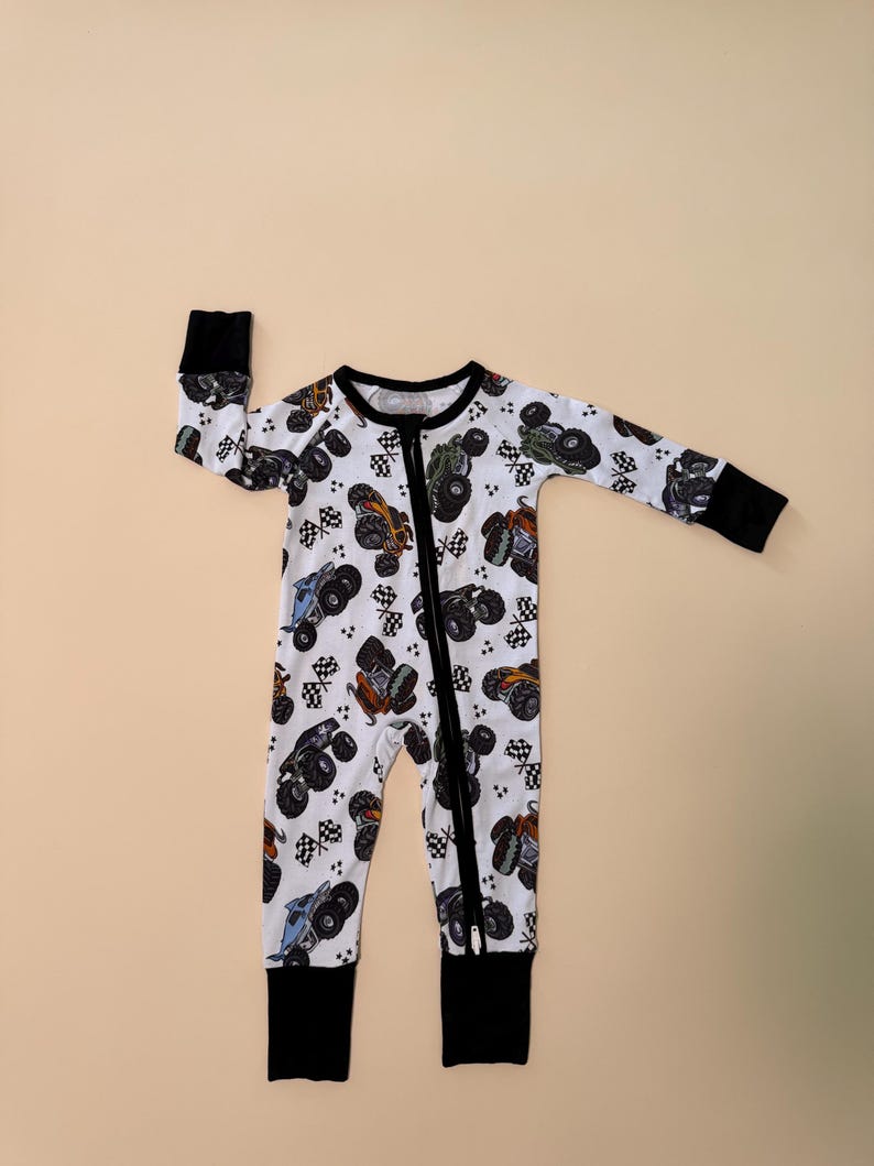 May include: White baby sleeper with a colorful monster truck pattern. The one-piece outfit features a black zipper, cuffs, and collar. The design includes monster trucks and checkered flags.
