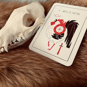Peut inclure: Une carte de tarot blanche avec une illustration de coq rouge et noir. La carte indique "X - Roue de la Fortune".