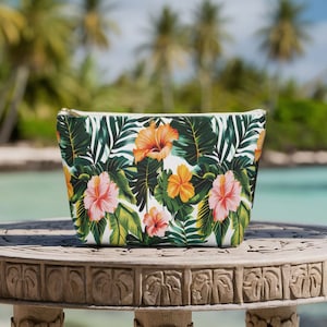 May include: A white zippered pouch with a tropical print featuring green leaves, pink and orange flowers. The pouch is sitting on a wooden table with a carved design.