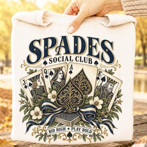 May include: A cream-colored sweatshirt featuring a graphic print with the text "SPADES SOCIAL CLUB" and "BID HIGH PLAY BOLD". The design includes illustrations of Jack, Queen, Ace, and King spade playing cards, floral accents, and a ribbon.