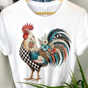 May include: A white short-sleeve t-shirt featuring a graphic of a rooster. The rooster has a black and white checkered pattern on its chest, a red comb, and a tail decorated with teal, orange, and leopard print feathers and floral accents.