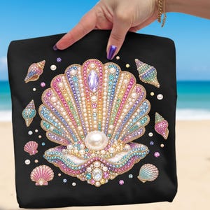 May include: Black tote bag featuring a large, ornate seashell design. The seashell is embellished with colorful gems and pearls, with smaller seashells and pearls scattered around. The design is set against a backdrop of a beach and ocean.