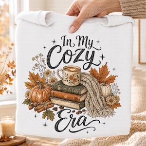 May include: A white sweatshirt featuring a graphic print with the text In My Cozy Era. The design includes a stack of three books, a floral mug, a pumpkin, cinnamon sticks, autumn leaves, and a knitted scarf with small star accents.