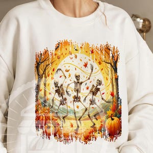 Puede incluir: Sudadera blanca con un diseño colorido de Halloween. El gráfico representa tres esqueletos bailando en una escena otoñal con calabazas, árboles y hojas cayendo. La obra de arte está sobre un fondo amarillo y naranja.