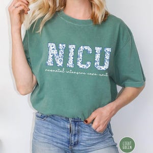 May include: A light green t-shirt with a white floral print that says "NICU" in large letters. The text below the letters says "neonatal intensive care unit".